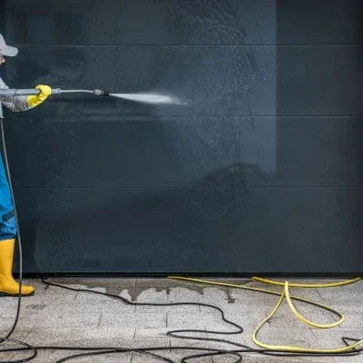 Worker pressure washing a dark gray paneled garage door with a wand, yellow hose on wet concrete driveway