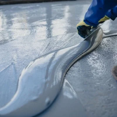 Worker in blue sleeves and yellow gloves spreading gray liquid resin coating across a concrete floor with a trowel