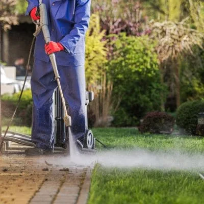 Worker pressure washing a brick paver walkway beside a green lawn with water mist